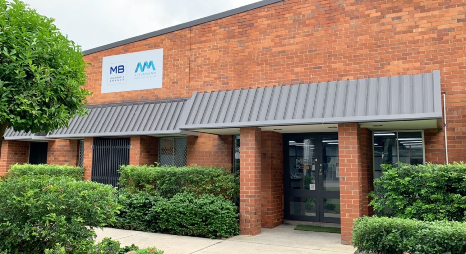 A brick building exterior with a gray metal canopy and a white sign reading Milners Brands and Aviva Mann, surrounded by green shrubs and trees.